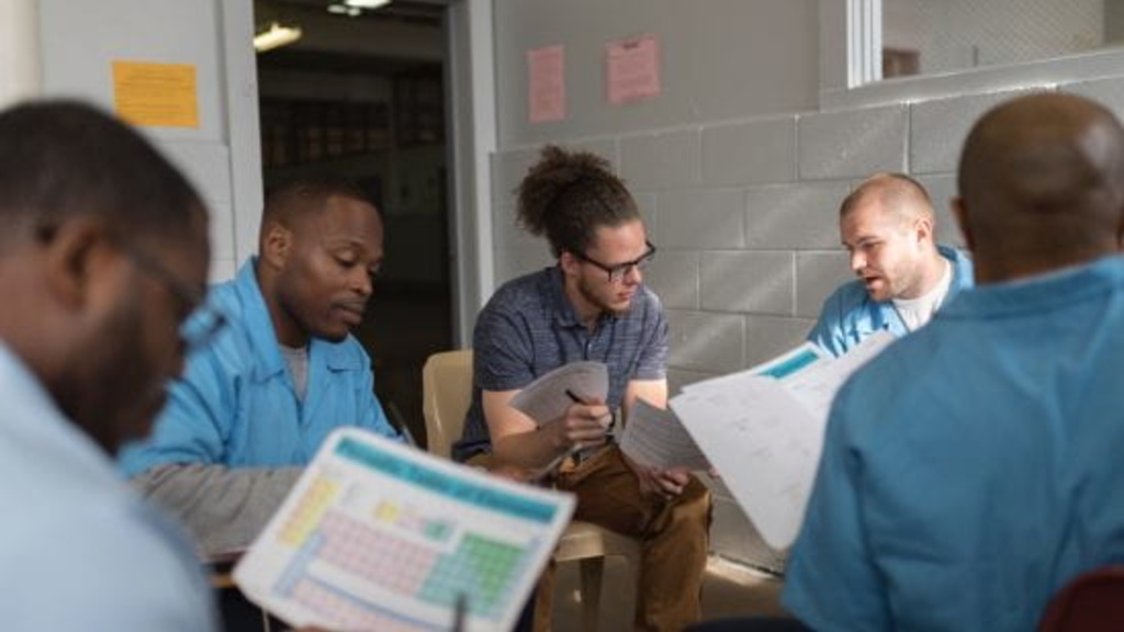 Men of different races sit around a table studying together. They are wearing matching blue shirts. 