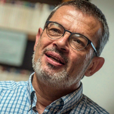 Headshot of smiling older man with grey short beard and glasses wearing a checked shirt. 