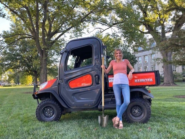 Woman standing against a small outdoor landscaping vehicle holding a shovel