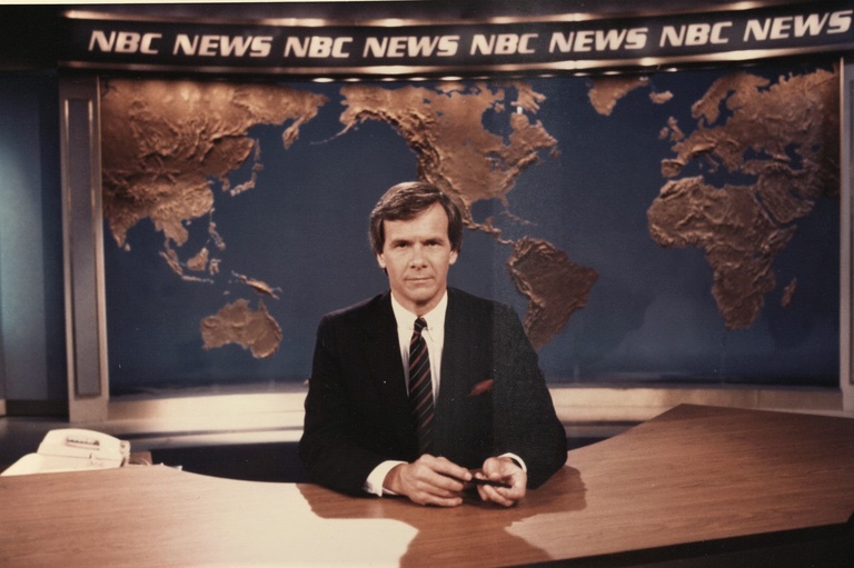 A man behind a desk with a large world map behind him and the words NBC News above him. 