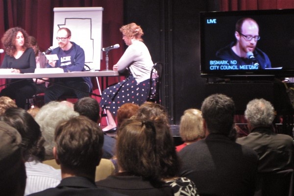 Three people are sitting at a meeting table with microphones, and to the right there is a projection of one of the people on a screen. An audience is in the foreground.