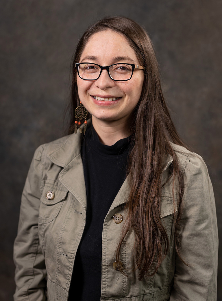 Headshot of young Latina wearing an olive blazer and black turtleneck. She has glasses and long brown hair. 