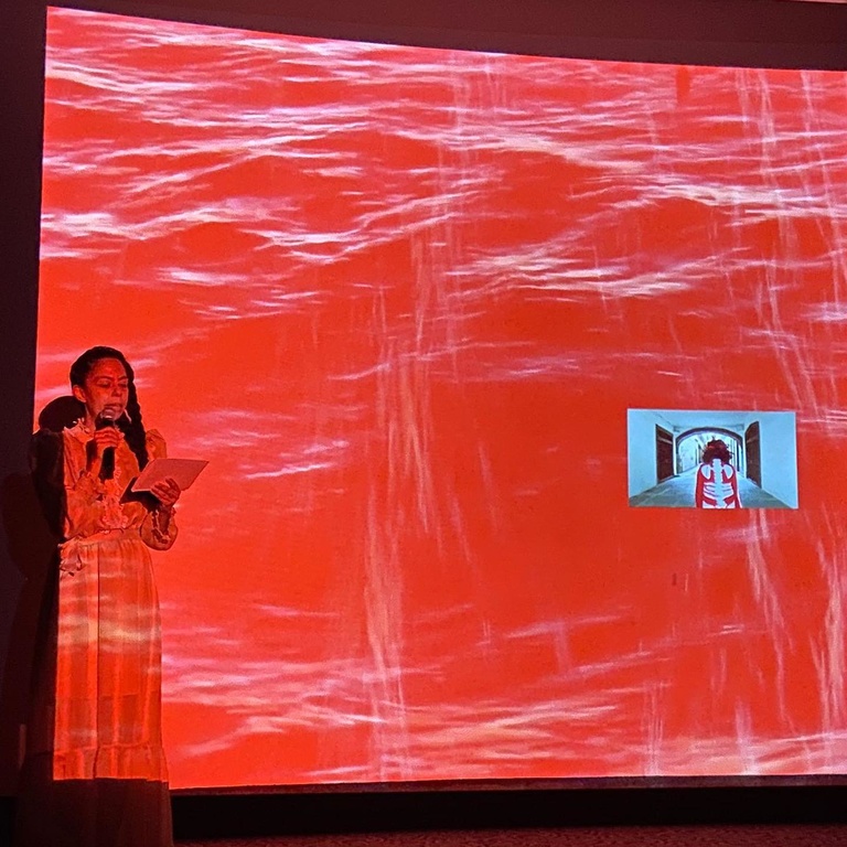 A women speaking in front of a red image with a woman in front of it.