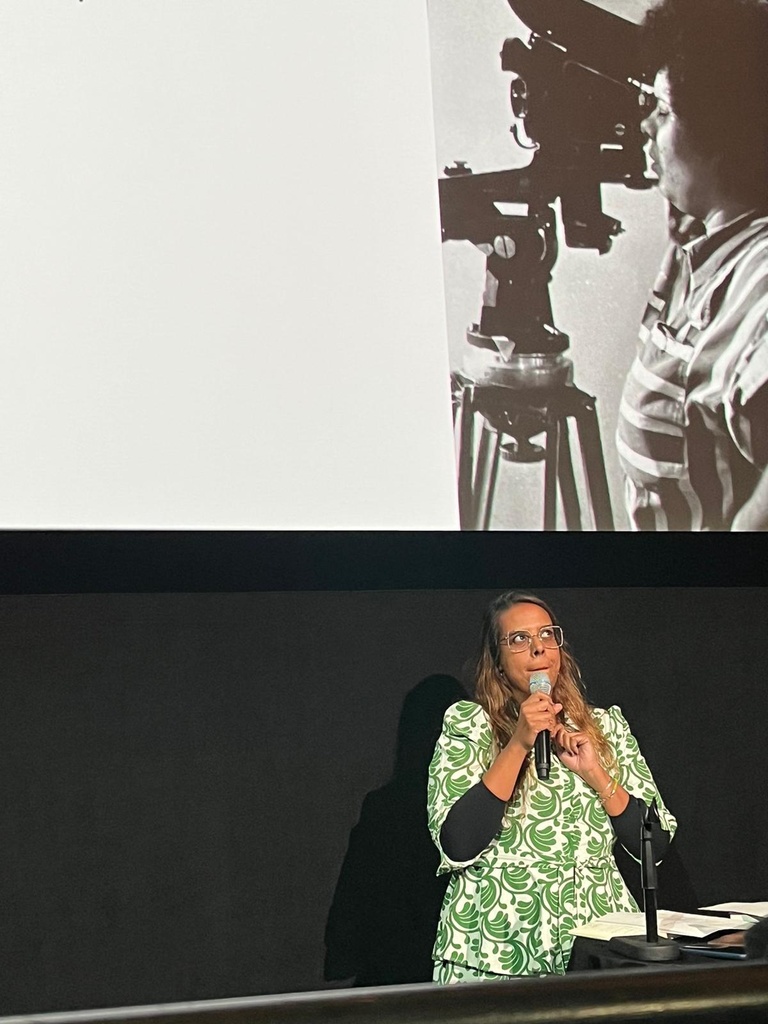A Black women speaking in front of an image with another black woman standing behind the camera