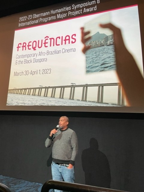 A balck man speaking in front of the Frequencias Poster on a screen