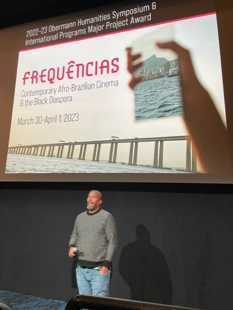 A black man in front of the Frequencias Poster on a screen