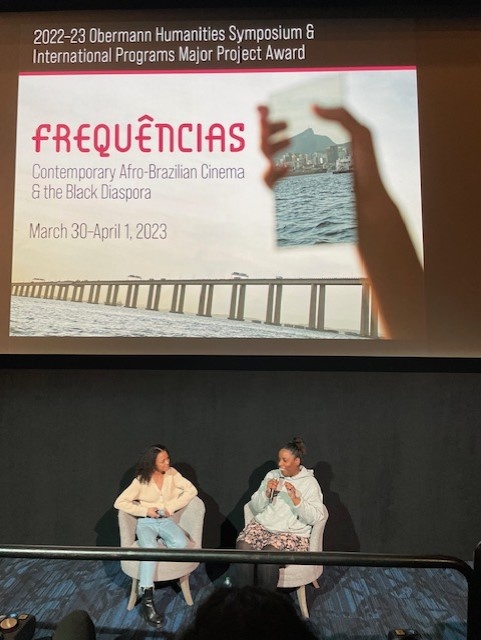 2 Black women speaking in front of the Frequencias Poster on a Screen