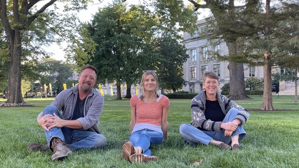 Three people sitting in a grassy area with trees behind them. 